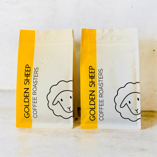 Two stand-up coffee roaster bags labeled GOLDEN SHEEP COFFEE ROASTERS, shown side by side on a light surface. Each bag has a white body with a bright yellow vertical band on the left and a simple black line drawing of a sheep on the right.