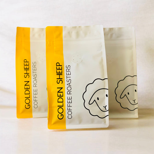 Three stand-up coffee roaster bags from Golden Sheep Coffee Roasters arranged in a row on a light neutral background. The bags are off-white with a bright yellow vertical band on the left side, featuring the brand name 'GOLDEN SHEEP COFFEE ROASTERS' in black text and a simple black line drawing of a sheep's head.