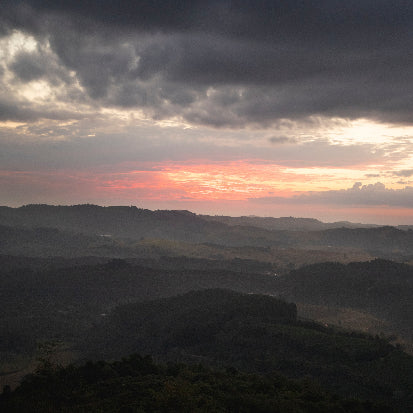 A scenic landscape photograph of rolling hills and mountains under a dramatic sunset sky with dark clouds, glowing orange light near the horizon, and misty atmospheric haze.