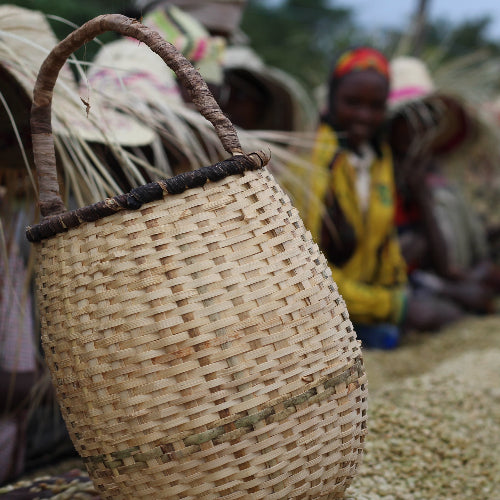 A close-up of a hand-woven natural bamboo basket with a dark brown handle, set against a blurred outdoor background showing people and other baskets.
