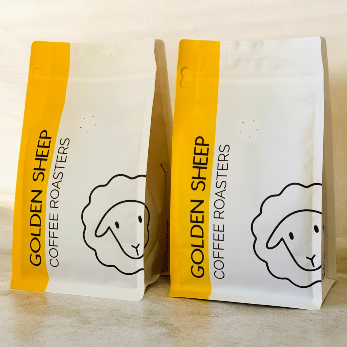 Two stand-up coffee bags from Golden Sheep Coffee Roasters on a light beige surface. The bags are white with a bright yellow vertical band on the left side, featuring the brand name 'GOLDEN SHEEP COFFEE ROASTERS' and a simple black line drawing of a sheep head.