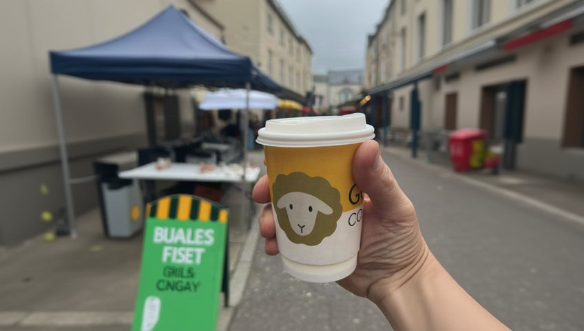 A hand holding a yellow and white coffee cup with a sheep logo, photographed on a blurred outdoor street market background.