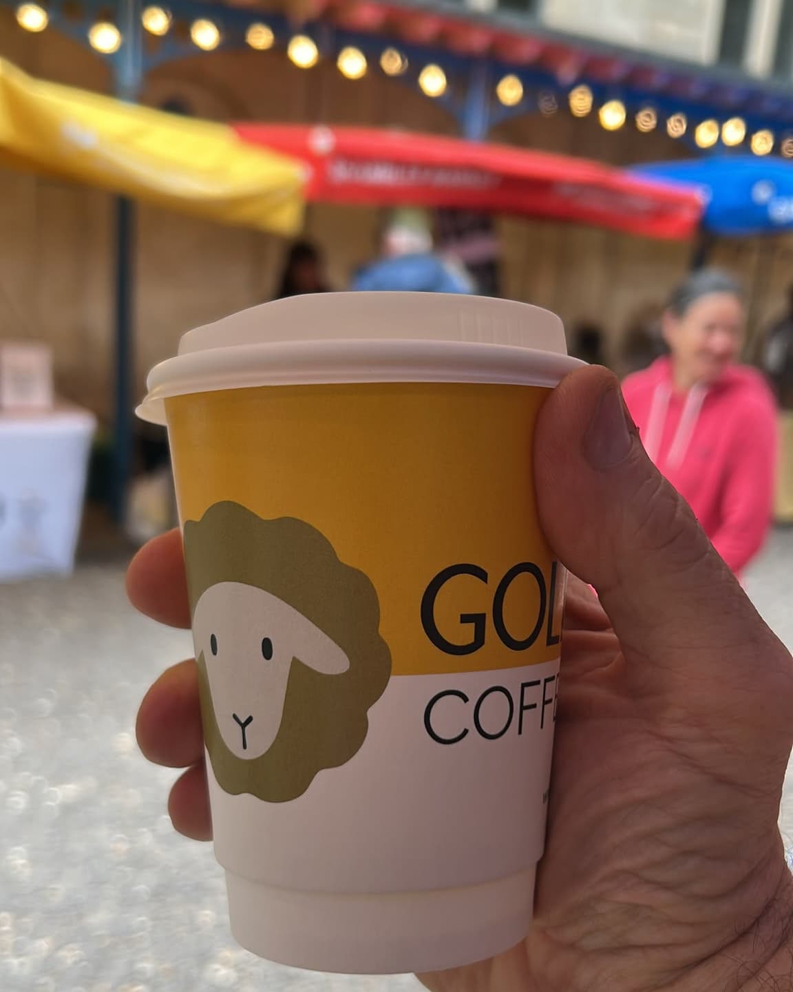 A hand holding a paper coffee cup with a yellow and white design, featuring a sheep logo and the partial text 'GOLD COFFEE', with a blurred outdoor market background.