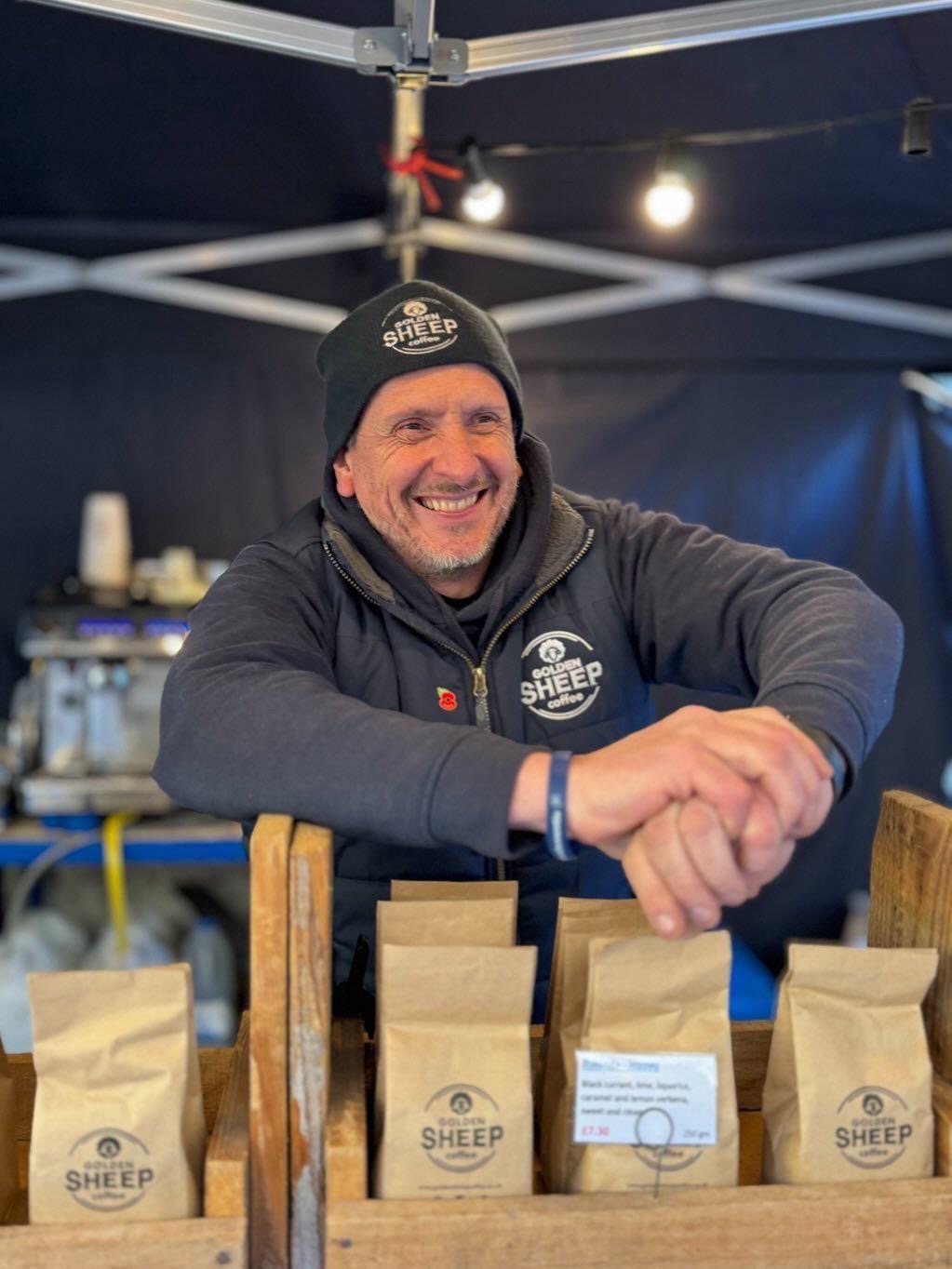 Man behind a counter with 'Sheep' branded coffee bags at an event.
