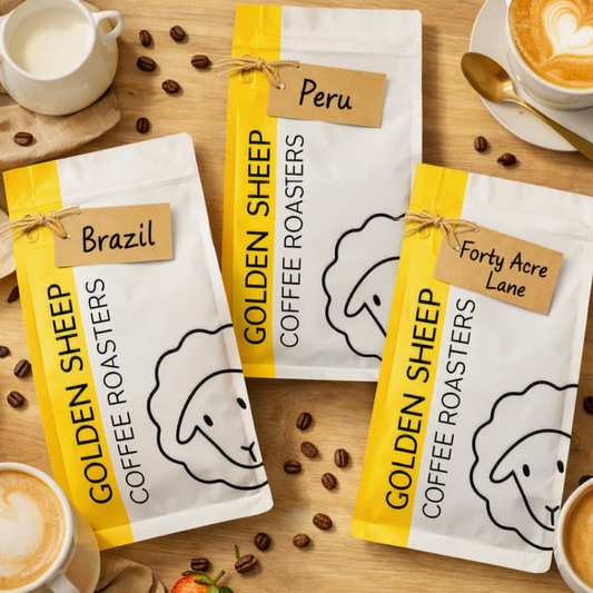 Three bags of Golden Sheep Coffee Roasters are arranged on a wooden table with coffee cups, a spoon, scattered coffee beans, and small food props. The bags are white with bright yellow vertical bands and kraft paper tags reading Brazil, Peru, and Forty Acre Lane.