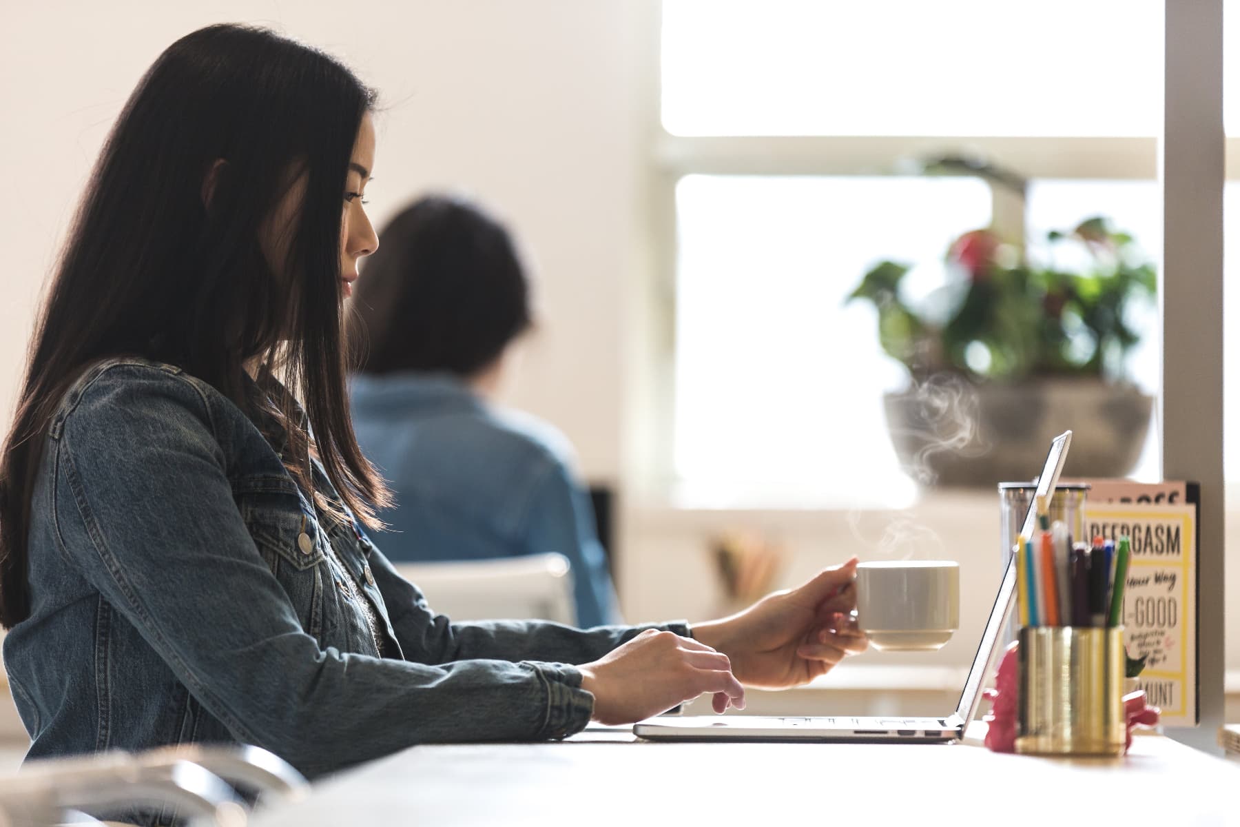 Woman using a laptop with a cup of coffee and stationery items on a desk.