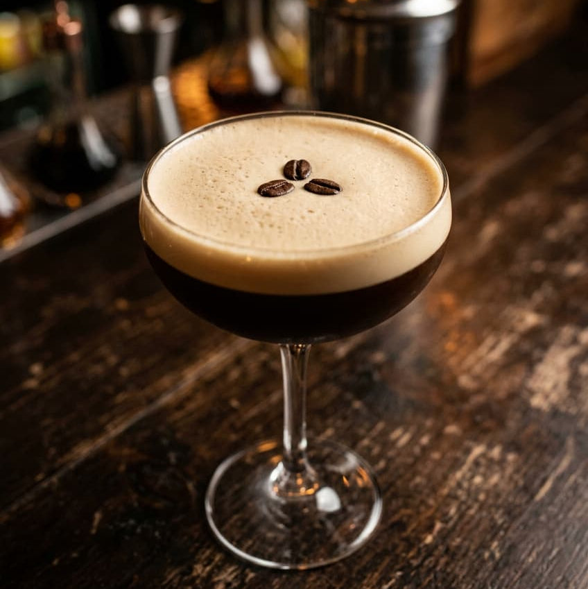 one espresso martini on a simple worktop, showing the foam and three-bean garnish clearly, with a cocktail shaker softly visible behind it. 
