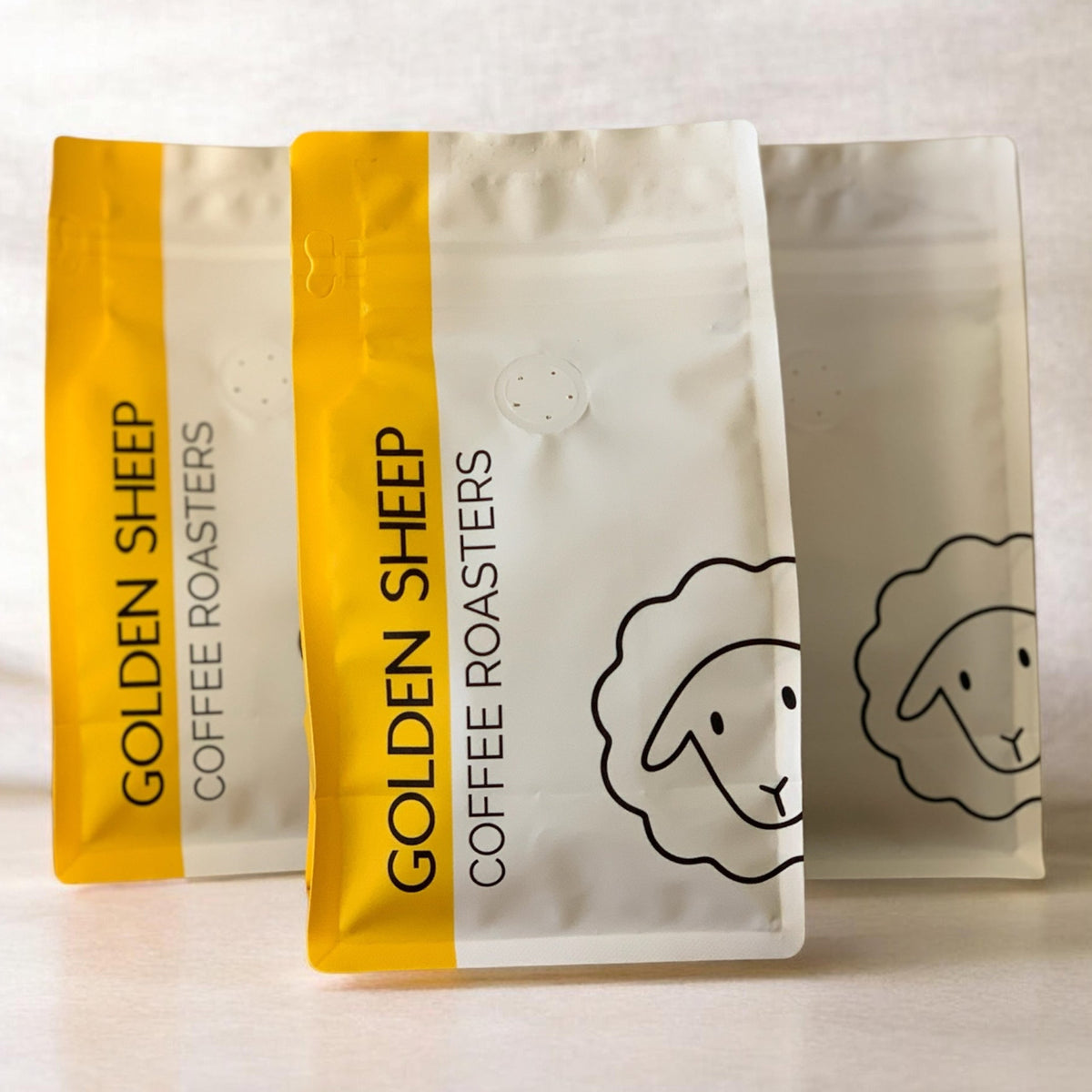 Three stand-up coffee roaster bags from Golden Sheep Coffee Roasters on a light neutral background. The front bag is white with a bold yellow vertical band and black text/logo, while the rear bags are partially visible in similar white and yellow packaging.