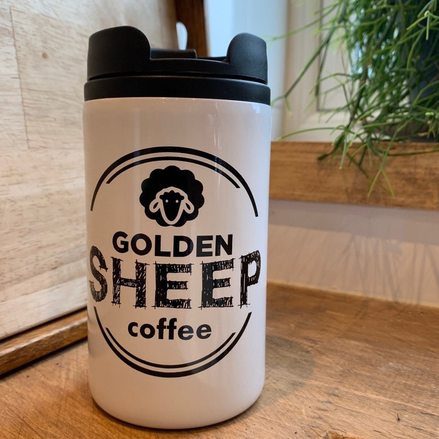 White tumbler with black lid and 'Golden Sheep Coffee' logo on a wooden surface.